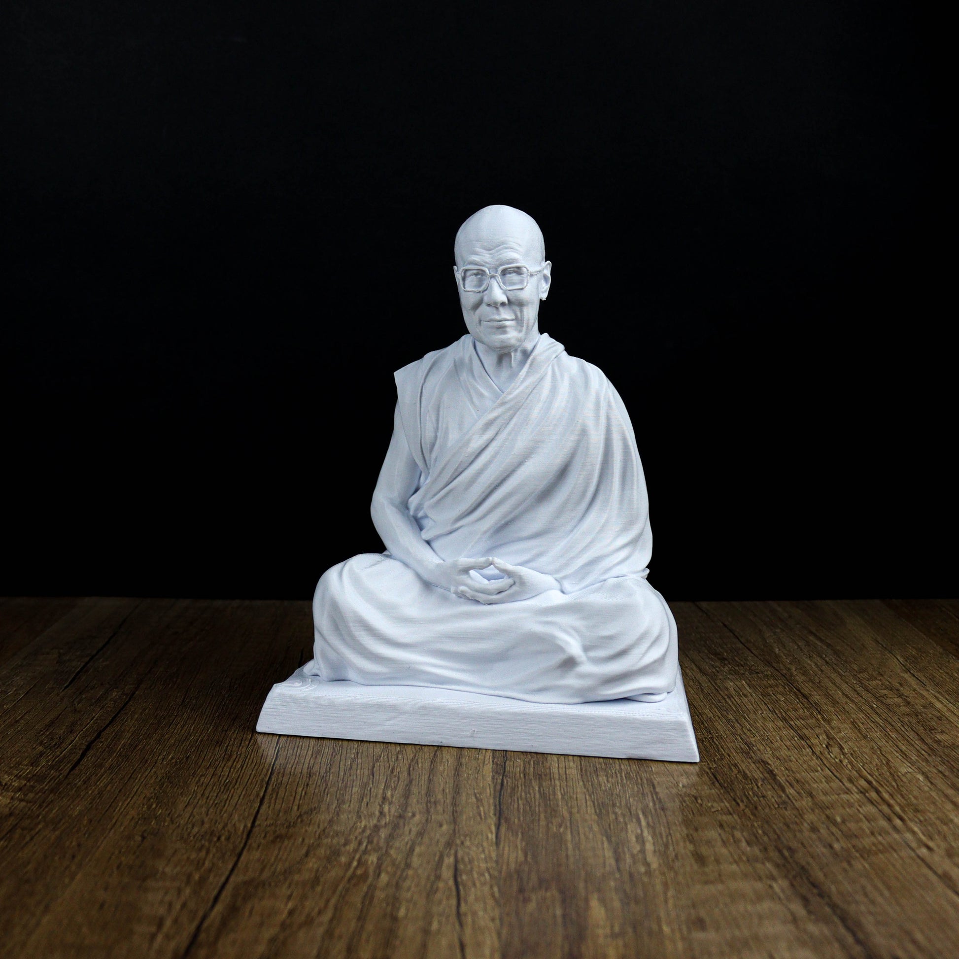 A white statue of a seated Buddha figure is displayed on a wooden surface against a black background.